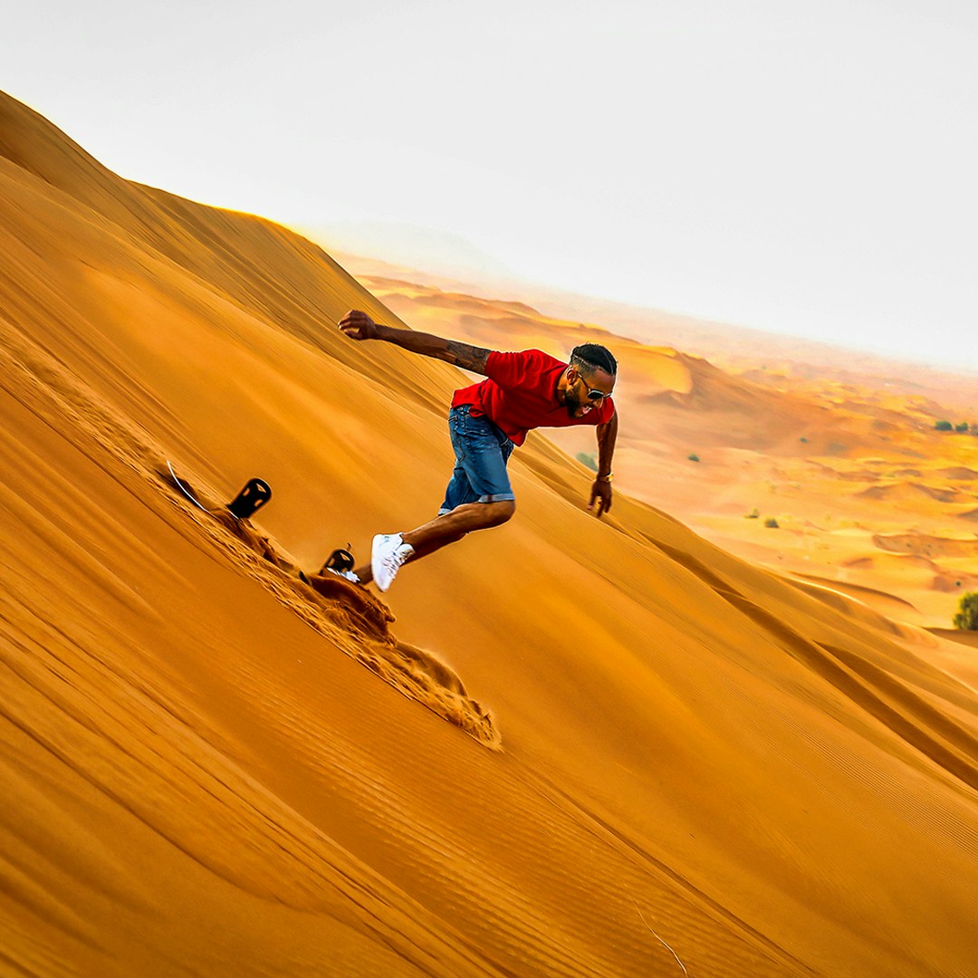 Person sandboarding down a desert dune during a 50 min quad biking, camel ride, and sandboarding tour.