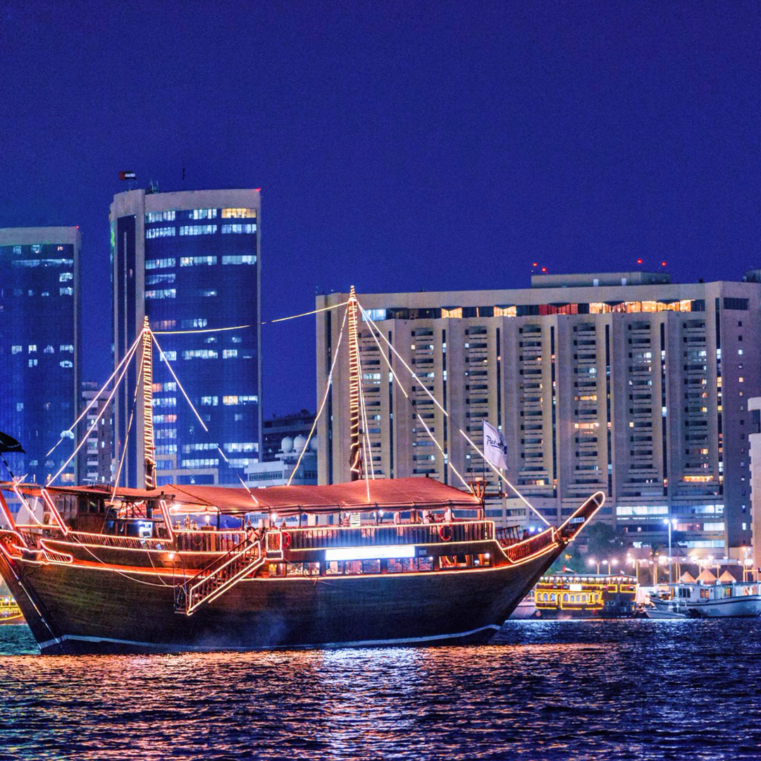 Dhow boat illuminated on Dubai Creek with city skyline at night.