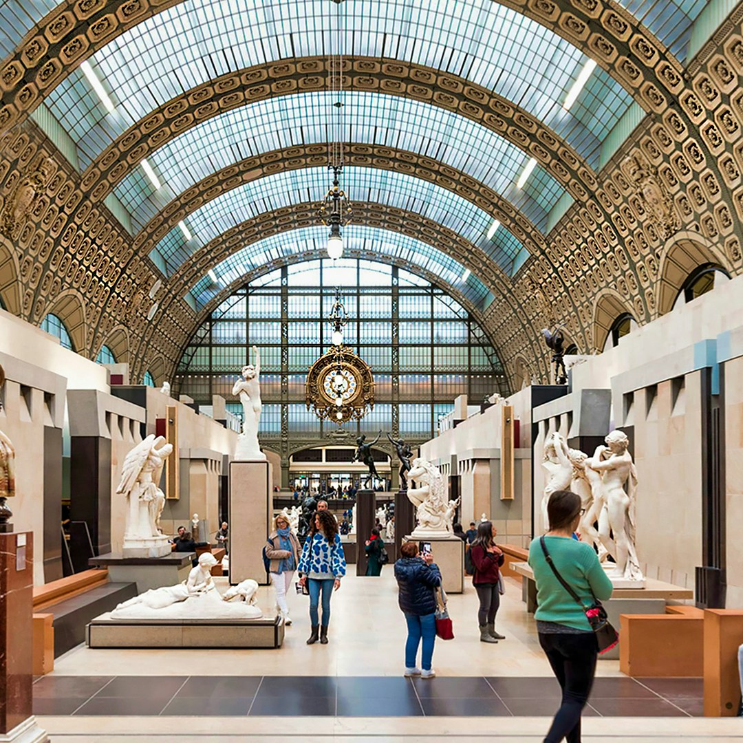Visitors exploring sculptures inside Orsay Museum, Paris, under ornate glass ceiling.